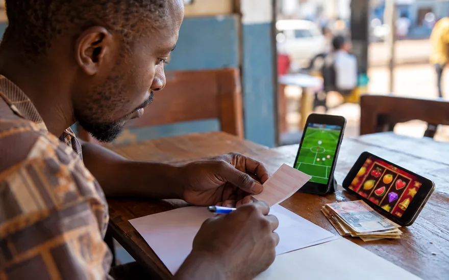 Zambia casino guide fact-checking scene with a physical betting slip and two blurred game phones in an adult Zambian setting.