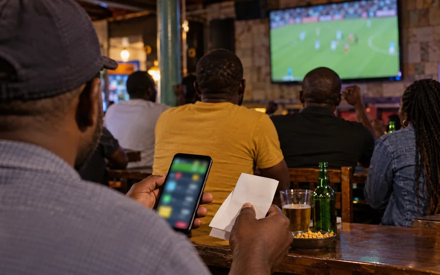 betPawa sports scene with adult Zambia supporters watching a football match beside a blurred betting phone.