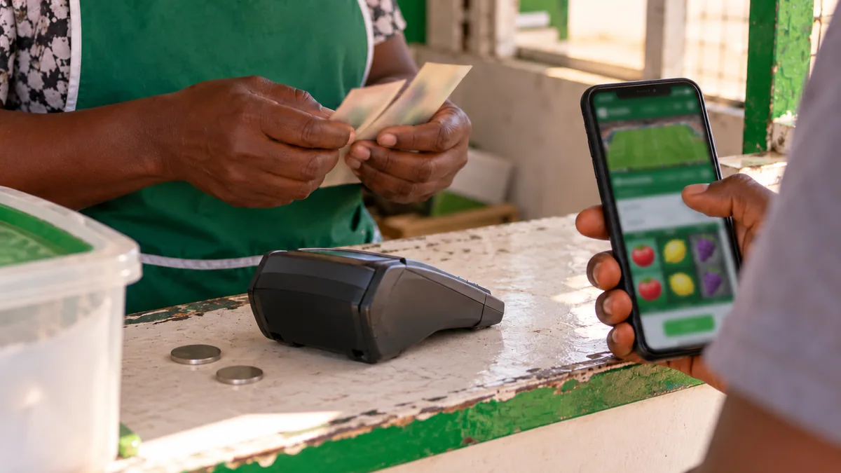 Cashier counting blurred banknotes beside a payment terminal, plain coins and a betting phone.